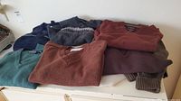 Eight sweaters folded on a white dresser in assorted colors: teal, rust, navy blue, gray with a pattern, burgundy, brown, purple, and light brown.