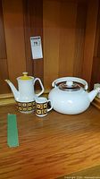 Photo of two sets of teapots and creamer on wooden surface. One set is Siena Ware with yellow, brown, and white geometric design, including a teapot and a creamer. The other item is a white ceramic teapot with a metal band around the lid.