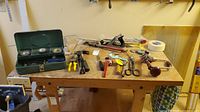 Top of workbench showing green metal toolbox with tapes and assorted hand tools scattered around including hammer, hand saw, pliers, scissors.