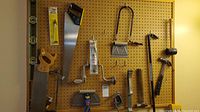 Wide view of pegboard wall with various hand tools including saws, hammers, level, and drilled bit sets hung and displayed.