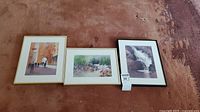 Three framed watercolor paintings by D Sykes laid on floor, showing scenes of street with people, groups in park, and waterfall.
