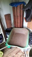 Wheelbarrow and garden fencing panels stored in a shed. Wheelbarrow is green metal with dirt inside. Wooden fencing panels lean against wall behind it.