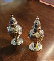 Pair of Victorian silver plated repousse salt and pepper shakers displayed on wood surface with visible floral embossed decoration and pierced lids.