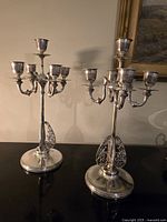 Photo of two silverplated five-light candelabras, standing upright, showing their ornate accent wings and polished round bases, placed against a beige wall.