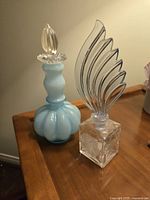 Two vintage perfume bottles on a wooden surface, one blue cameo glass with bulbous body and clear stopper, one clear square block glass with curved Art Nouveau stopper.