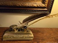 Photo showing antique brass double inkwell with two open inkwell openings, decorated with ornate engravings and patina, two feathers placed in the compartments (not included in the lot).