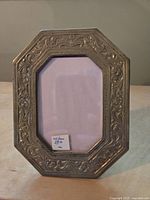 Photo of one brass frame showing octagonal shape, embossed floral decoration, glass front, and approximate size of 5 by 4.5 inches.