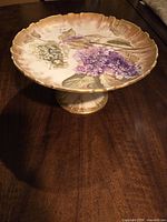 Photo showing the overall shape and height of the china comport with lilac floral decoration and gold gilded edges on a wooden surface.