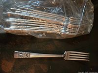 Forks in plastic bag with one fork out showing ornate crest handle design and four tines.