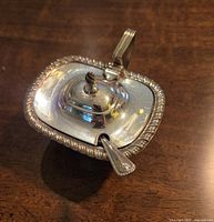 Closed mustard dish showing gadroon edge and hinged lid with finial on a wooden surface.