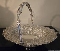 Side angle view of silver hand-hammered repoussé cake basket showing detailed floral and scroll design on basket and handle