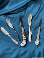 Five mixed silver plated butter and cheese cutting utensils displayed on blue cloth background. Various ornate embossed handles and blades, including one with cloisonne floral handle.