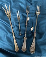 Four silver plated pickle forks with diverse handle styles showing details of tines and handles against a blue satin background.