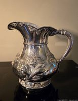 Front view of ornate silverplated water jug showing repousse decoration, handle and scalloped lip