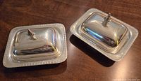 Pair of silver plated rectangular butter dishes with covers and decorative spoke finials, on a wooden surface.