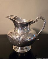 Photo shows a silverplate water jug with bulbous body, embossed decorative elements, a flared spout, and an ornate scroll handle. Surface is reflective but has minor scratches visible on one side.