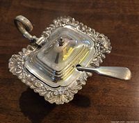 Silver-toned mustard dish with intricate scrolled edges and matching spoon on wood surface, lid closed