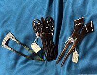 Three hair combs displayed on blue fabric including a silver-tone metal comb, and two dark brown plastic combs, one with rhinestones.