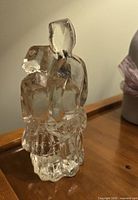 Close-up front view of the clear glass sculpture depicting two abstract figures embracing, placed on a wooden surface near a vase with ribbon.