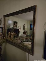 Photo showing the large rectangular mirror mounted on the wall reflecting a living room with furniture and paintings. The gold carved frame details are visible.