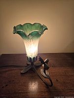 Lamp lit showing the green and white gradient tulip-shaped glass shade and detailed amber brass bird base on a wooden surface.