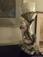 Wood nymph sculpture holding a flower-shaped candleholder with a textured glass bowl on top, placed on a dark surface with paintings in the background.