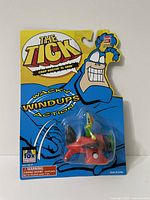 Front view of sealed package showing The Tick branding and visible Crop Dustin El Seed figure on red airplane toy inside clear blister packaging.
