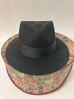 Black Biltmore Harrison fedora hat displayed on a vintage-style hatbox cover with postage stamp designs, showing front and side views of the hat.