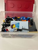 Plastic storage bin filled with assorted loose Lego bricks and pieces in multiple colors, including black, white, blue, red, gray and yellow. Red lid seen in background.