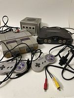 Front and top view of Nintendo 64, Super Nintendo Entertainment System, and Nintendo GameCube consoles with attached controllers and cables on white background.