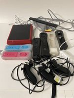 Photo showing the red Nintendo Wii Mini console along with multiple remotes in black, blue, and pink, some with silicone covers and wrist straps. Also includes a sensor bar and various cables.