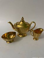Full set front view showing teapot, creamer, and bowl with gold reflective finish and floral etching.