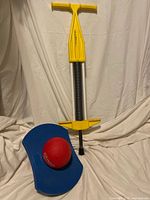 Yellow Pogo-Roo pogo stick with metal spring and T-handle shown upright beside the blue curved Air Pogo jumper platform with red ball resting on it.