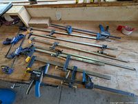 Image showing about 8 bar clamps on a workbench, various sizes and types, mostly metal with blue and yellow handles, quick-release style clamps visible.