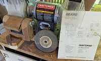 Front view of Craftsman bench grinder showing the coarse grinding wheel, wooden grinding guides, and original owner's manual placed beside it.