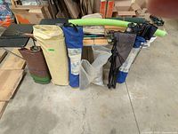 Camping items packed in bags and cases on and near camp tables, including tent poles, chairs, and fishing rods.