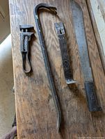 Photo shows four items on a wooden surface: adjustable wrench, crow bar, pry bar, and machete with black riveted handle.