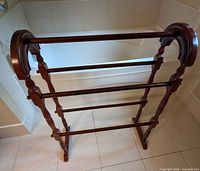 Front view of wooden quilt stand, showing three horizontal slats and curved top ends, placed on tiled floor near bathtub.