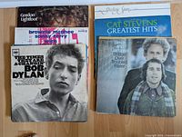 Twelve folk music LP records on a wooden floor showing front covers including Bob Dylan, Gordon Lightfoot, Brownie McGhee & Sonny Terry, Steeleye Span, Cat Stevens Greatest Hits, and Simon & Garfunkel Bridge Over Troubled Water.