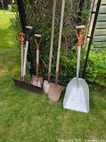 Five garden shovels and spades standing on grass in front of a black metal trellis and greenery in the background. Tools vary in size and blade shape with wooden and plastic handles.