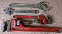 Four large adjustable wrenches arranged side by side on a carpeted floor including two red pipe wrenches and two silver crescent wrenches.