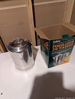 Stainless steel coffee percolator next to its original box on a white surface, highlighting the pot and packaging.