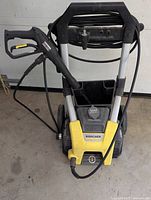 Full view of Karcher electric pressure washer with yellow and black color, showing frame, wheels, soap dispenser, hose, and trigger gun.