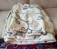 Folded white fabric duvet cover with intricate embroidered floral vine patterns in beige and grey tones on a soft fabric, folded and placed on couch.
