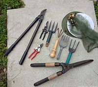 Assorted small garden tools laid out on concrete surface including large shear, several hand pruning secateurs, cultivators, forks, and insect net.