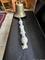 Solid brass bell laid on a black surface showing the brass body and attached clanger cord made of thick knotted rope with monkey's fist knots.