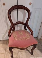Front view showing carved wooden frame with oval backrest with berry and leaf motif and pink floral tapestry seat.