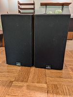 Front view of two black grille Celestion bookshelf speakers side by side on a wooden table with visible wood grain surface.