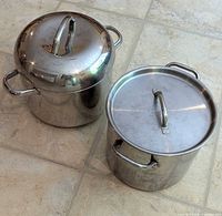 Two large stainless steel pans side by side on tile floor, both with lids, one pan is taller with a dome lid handle.