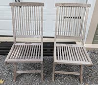 Front view of two teak folding chairs showing vertical slatted seat and backrest, no arms, weathered teak wood finish.